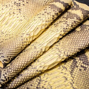 May include: A folded sheet of textured material featuring a repeating scale pattern in shades of pale yellow and dark brown. The surface displays a natural, organic appearance with overlapping, diamond-shaped scales that create a tactile, layered effect.