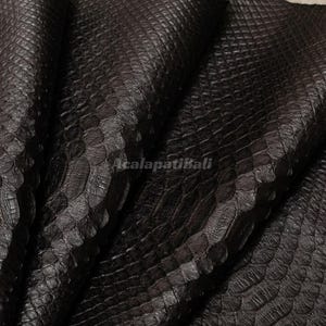May include: A dark brown, textured material featuring a repeating, overlapping scale pattern across the entire surface. The material is folded, creating deep shadows and highlights. The text AcalapatiBali is printed in the centre of the image.