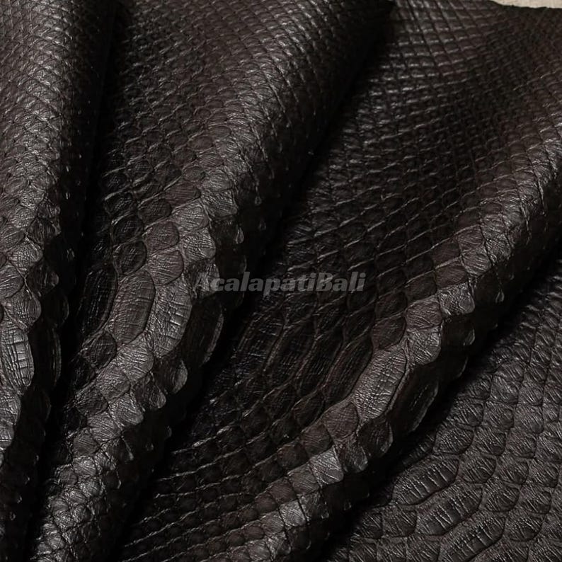 May include: A dark brown, textured material featuring a repeating, overlapping scale pattern across the entire surface. The material is folded, creating deep shadows and highlights. The text AcalapatiBali is printed in the centre of the image.