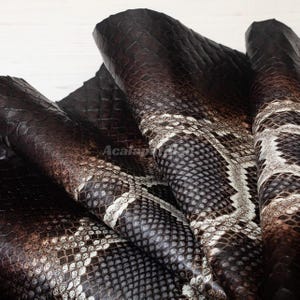 May include: A piece of dark brown and cream-coloured snakeskin leather with a distinct, repeating scale pattern. The material is folded, showing a textured surface with deep brown tones transitioning into lighter, off-white scale details throughout.