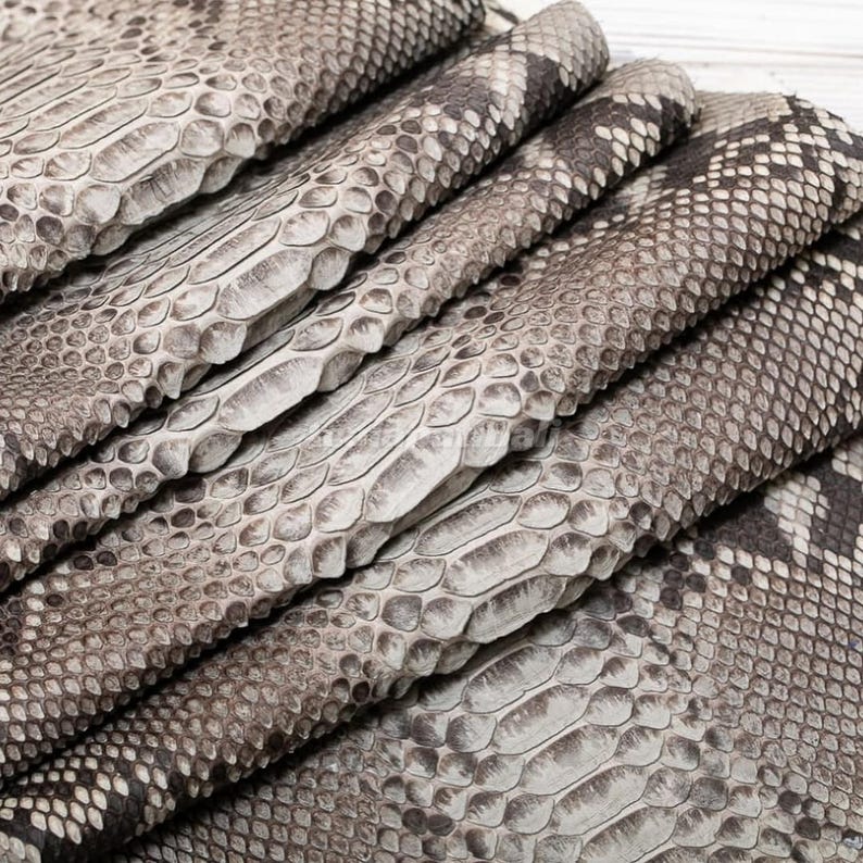 May include: A folded piece of textured snakeskin leather featuring a natural pattern of overlapping scales in shades of gray, beige, and off-white. The material displays a distinct reptilian texture with varying scale sizes across the surface.