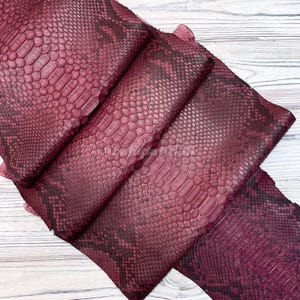 Può includere: Un pezzo di pelle di serpente color bordeaux caratterizzato da un distinto motivo a scaglie in rilievo con segni più scuri nei toni del nero. Il materiale è piegato in più strati, mettendo in risalto la superficie testurizzata e il colore rosso.