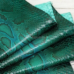 Peut inclure: Un morceau de cuir de serpent vert émeraude présentant un motif d'écailles répétitif et distinct. Le matériau affiche une teinte verte vive et profonde avec des contours d'écailles plus sombres, créant un aspect reptilien texturé.