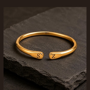 May include: A gold-toned open cuff ring featuring a smooth, polished finish. The two ends of the ring are flattened and each displays a single engraved letter, with the letter S on one side and the letter Z on the other side of the opening.