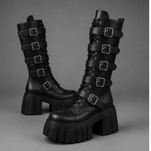 May include: A pair of black knee-high boots featuring a thick platform lug sole and a chunky block heel. The boots have a lace-up front closure and five horizontal straps across the shaft, each secured with a silver-toned metal buckle.