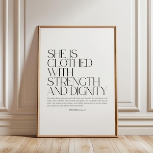 Peut inclure: Une affiche encadrée rectangulaire avec du texte noir sur fond blanc. Le texte indique : SHE IS CLOTHED WITH STRENGTH AND DIGNITY, suivi du texte complet de Proverbes 31:24-27. Le cadre est en bois clair avec un profil fin et épuré.