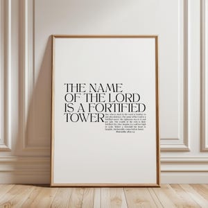 Peut inclure: Une affiche murale rectangulaire verticale dans un cadre en bois clair avec du texte noir sur fond blanc. Le texte indique : THE NAME OF THE LORD IS A FORTIFIED TOWER, suivi du texte complet de Proverbes 18:9-12 dans une police plus petite.