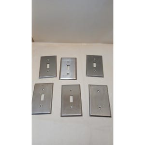 May include: A set of six rectangular brushed metal light switch plates, each featuring a central vertical toggle opening and two screw holes positioned at the top and bottom, arranged in two rows of three on a neutral surface.