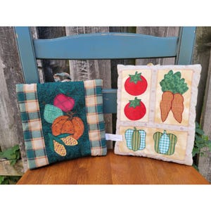 May include: Two square fabric potholders featuring quilted vegetable designs. The left one has a dark green center with an apple, pear, pumpkin, and corn. The right one has a cream background with two red tomatoes, two carrots, and two peppers.