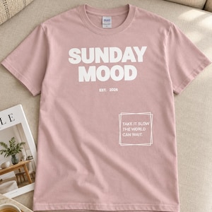 Könnte beinhalten: Ein altrosa T-Shirt mit kurzen Ärmeln und dem weißen Schriftzug SUNDAY MOOD sowie EST. 2026 auf der Brust. Darunter befindet sich ein weißes Quadrat mit dem Text TAKE IT SLOW THE WORLD CAN WAIT. Das T-Shirt hat einen Rundhalsausschnitt.