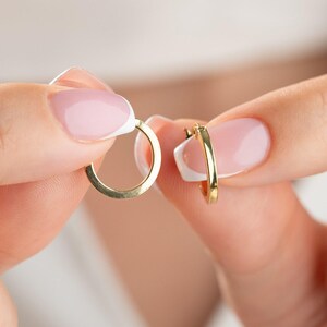 May include: A pair of small, polished gold hoop earrings with a smooth, circular design. Each earring features a simple, continuous band with a hinged closure mechanism, showcasing a bright, reflective metallic finish and a classic, minimalist shape.