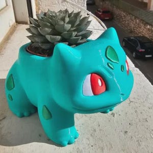 May include: A teal-colored planter shaped like the character Bulbasaur, featuring large red eyes, white pupils, and dark green spots on its back and legs. A green succulent plant is potted inside the hollow opening on the creature's back.
