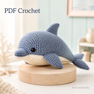 Puede incluir: Un juguete de delfín tejido a ganchillo en azul y crema con ojos de plástico negro. El texto PDF Crochet es visible en la esquina superior izquierda, y el texto AmiLoomStudio es visible en la esquina inferior derecha de este peluche hecho a mano.