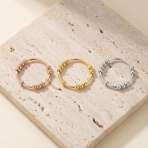 Op de afbeelding: Drie dunne metalen ringen, één in roségoud, één in geelgoud en één in zilver, elk voorzien van een reeks kleine, beweegbare bolvormige kralen langs de band, ontworpen om rond het cirkelvormige draadframe van de ring te draaien.