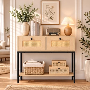 May include: A light wood console table with two drawers featuring cane webbing panels and black knobs, supported by a black metal frame. The table includes a lower shelf holding a woven basket and two rectangular cane-accented storage boxes.