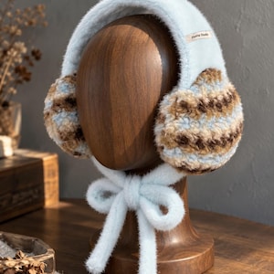 May include: Light blue fuzzy earmuffs featuring a brown, tan, and light blue striped pattern on the ear covers. The headband has a soft texture and includes a small tag labeled Haena Studio. Two long, light blue fuzzy ties hang down.