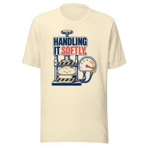 May include: A cream-colored short-sleeve t-shirt featuring a graphic of a hydraulic press compressing a small, smiling dumpling. The text reads "HANDLING IT SOFTLY" in dark blue and red, with a pressure gauge indicating "SOFT" in red.