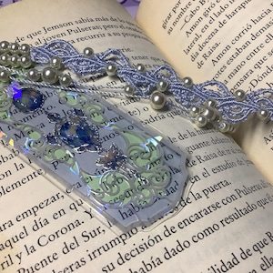Puede incluir: Un marcapáginas rectangular con acabado holográfico en tonos verde claro y azul, que incluye un detalle central similar a una gema azul. Un cordón trenzado de color violeta claro con perlas blancas está sujeto a la parte superior.