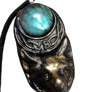 May include: A pendant featuring a sculpted bronze-colored fox head at the base, topped with a crescent-shaped frame containing an iridescent blue-green labradorite gemstone. The piece is suspended from a simple black cord with a silver-toned bail.