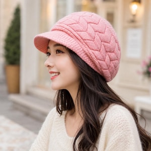 May include: A soft pink knit cap featuring a textured chevron pattern and a short, stiff brim. The hat is constructed from a thick, warm yarn with a visible woven stitch design that covers the crown and sides, providing a structured fit.