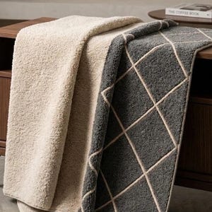 May include: A reversible throw blanket featuring a textured cream-colored side and a dark gray side with a white diamond grid pattern. The blanket is draped over a wooden surface, showcasing its thick, plush material and finished edges.