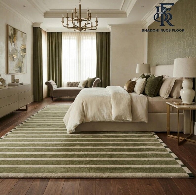 May include: A large rectangular area rug featuring a repeating pattern of wide olive green and narrow cream horizontal stripes. The rug is positioned on a dark wood floor in a bedroom setting, extending from the foreground toward a bed and chaise lounge.