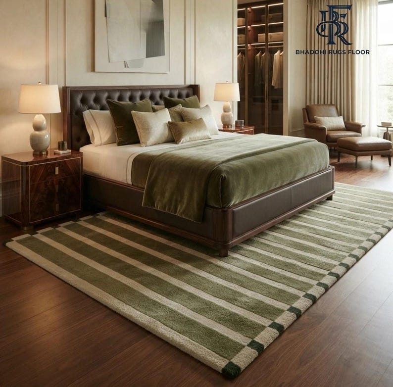 May include: A rectangular area rug featuring a striped pattern in alternating shades of olive green and cream. The rug is positioned on a dark wood floor beneath a bed with a dark brown headboard, two bedside lamps, and matching bedding.