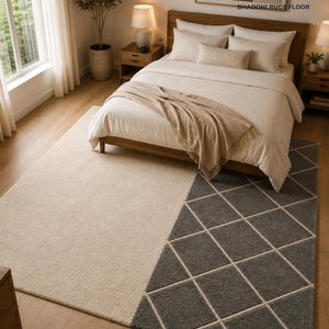 May include: A rectangular area rug featuring a split design with a solid cream-colored section and a dark gray section patterned with a white diamond grid. The rug is positioned on a wooden floor beneath a bed with cream bedding and a tan throw.