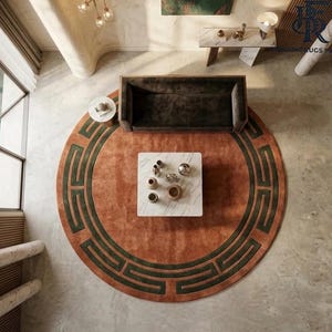 May include: A large circular area rug featuring a burnt orange base color with a dark green geometric Greek key border pattern. The rug is positioned under a dark brown rectangular sofa and a square white marble coffee table with decorative items.