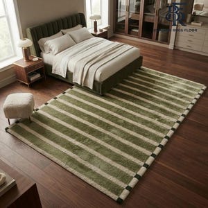 May include: A rectangular area rug featuring a pattern of wide olive green stripes alternating with narrower cream-coloured stripes. The rug has a dark green border with small black square accents along the edges, placed on a dark wood floor.