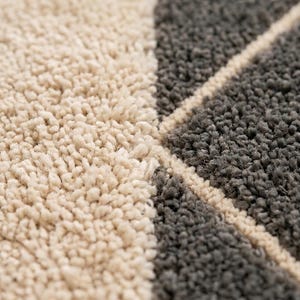 May include: A high-pile area rug featuring a geometric pattern of cream and dark gray tufted sections separated by thin cream lines. The top right corner displays a logo with the text BHADOHI RUGS FLOOR and a stylized monogram symbol.