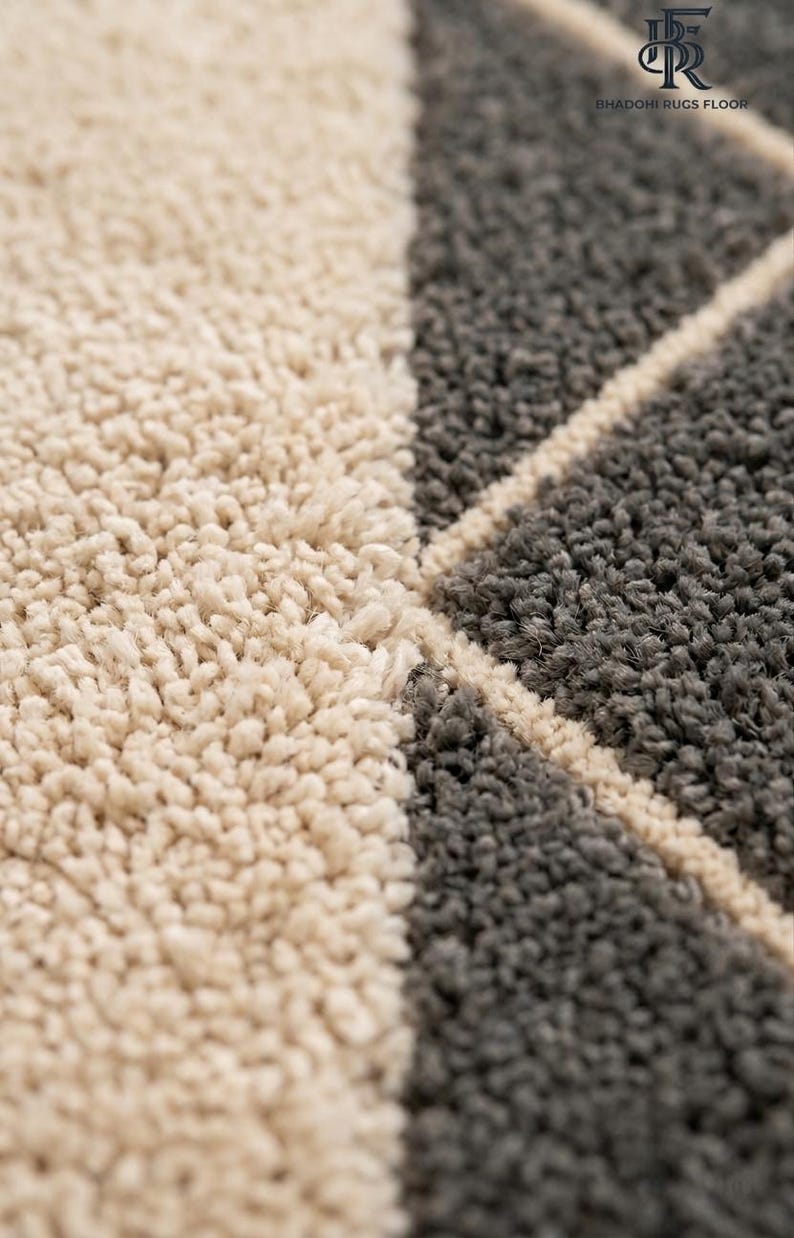 May include: A high-pile area rug featuring a geometric pattern of cream and dark gray tufted sections separated by thin cream lines. The top right corner displays a logo with the text BHADOHI RUGS FLOOR and a stylized monogram symbol.