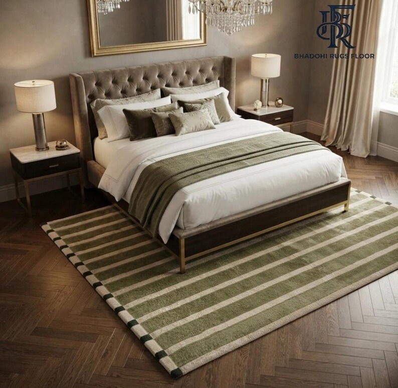 May include: A rectangular area rug featuring a pattern of alternating olive green and cream-coloured horizontal stripes. The rug is positioned beneath a tufted upholstered bed frame with white bedding, two nightstands with lamps, and a chandelier.