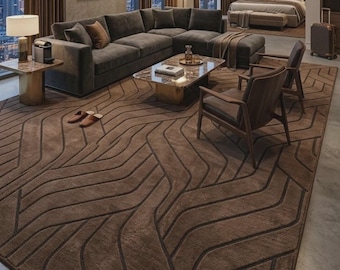 May include: A large rectangular area rug featuring a deep brown colour palette with a repeating pattern of dark, recessed, flowing geometric lines. The rug covers a significant portion of the floor in a room with a sofa, chairs, and a coffee table.