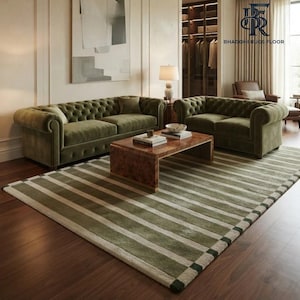 May include: A large area rug featuring a pattern of wide olive green and cream-coloured horizontal stripes. The rug is placed on a dark wood floor beneath a wooden coffee table, two tufted olive green velvet sofas, and a table lamp.