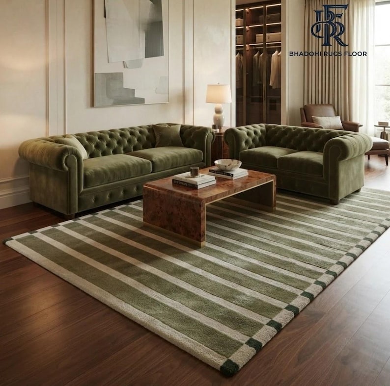 May include: A large area rug featuring a pattern of wide olive green and cream-coloured horizontal stripes. The rug is placed on a dark wood floor beneath a wooden coffee table, two tufted olive green velvet sofas, and a table lamp.