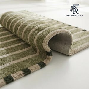 May include: A rectangular rug featuring a striped pattern in shades of sage green and cream. The rug is partially folded, revealing a textured underside and a border with dark green and cream squares. The logo BHADOHI RUGS FLOOR is visible.