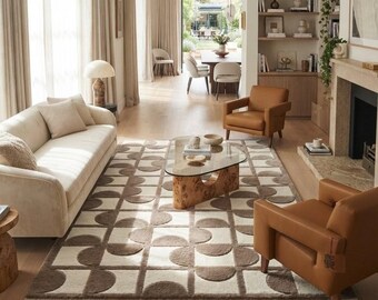 May include: A rectangular area rug featuring a repeating geometric pattern of brown semi-circles on a cream background. The rug is positioned in a living room with a cream sofa, two tan leather armchairs, and a glass-topped coffee table with a wooden base.