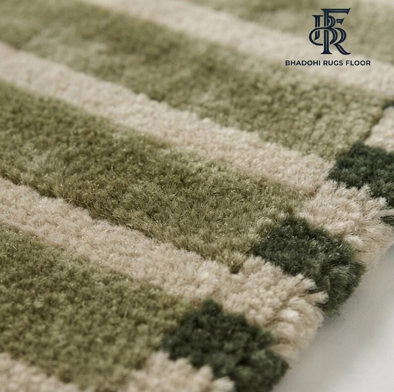 May include: A tufted rug featuring a repeating pattern of horizontal stripes in shades of sage green, olive green, and cream. The surface has a thick, textured pile. The top right corner displays the logo and text BHADOHI RUGS FLOOR.
