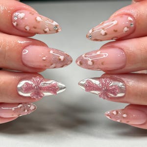Peut inclure: Ongles en amande avec une base nude brillante ornée de motifs léopard marron et blanc, de petits strass transparents et d'un emblème en forme de croix tridimensionnelle en paillettes argentées et roses sur les annulaires.