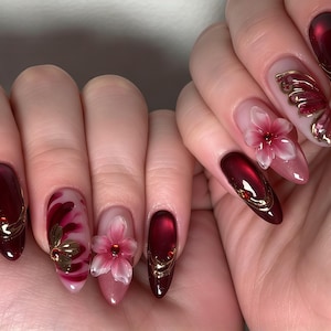 May include: Almond-shaped nails featuring a deep burgundy red polish with a glossy finish. The design includes three-dimensional pink floral accents, gold metallic line detailing, and small red rhinestones embedded on select nails.