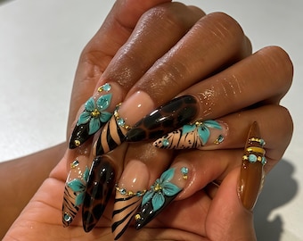 May include: Stiletto-shaped nails featuring a mix of designs including brown leopard print, black tiger stripes, and nude bases. The nails are decorated with three-dimensional light blue floral accents, small gold beads, and tiny light blue rhinestones.