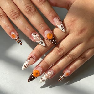 May include: Almond-shaped nails featuring a clear base with intricate autumn-themed nail art. Designs include brown and white polka dots, horizontal stripes, white swirls, bows, and small orange 3D pumpkins accented with tiny white pearls.