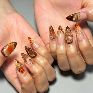 Puede incluir: Uñas en forma de almendra con un patrón de carey en tonos marrones y naranjas, acentuadas con dijes metálicos dorados, pequeñas perlas y detalles intrincados de alambre. El diseño incorpora secciones transparentes y motivos florales en relieve 3D.