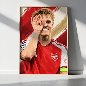 May include: A framed poster featuring a portrait of a soccer player wearing a red and white Arsenal jersey with the team crest and a Champions League patch. The player is smiling and making an eye gesture with one hand against a red brushstroke background.