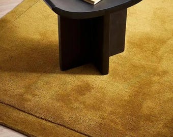 Puede incluir: Un taburete negro curvado con base en forma de cruz sobre una alfombra texturizada de color amarillo mostaza. Un pequeño libro de color crema y un porta-incienso de madera marrón se encuentran sobre el asiento de acabado mate.