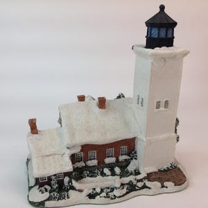 May include: A miniature lighthouse figurine featuring a tall white tower with a black lantern room and a red brick building attached. The structure is covered in white snow, with small green bushes at the base and text inscribed on the front corner.
