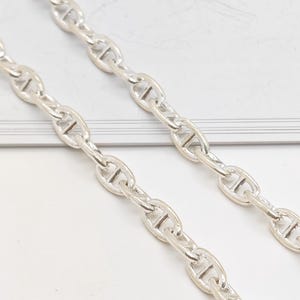 Op de afbeelding: Een zilverkleurige metalen ketting met ovale schakels, elk voorzien van een horizontale dwarsbalk in het midden. De ketting bestaat uit gepolijste, onderling verbonden schakels die een uniform, herhalend patroon vormen over de gehele lengte.