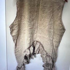 May include: A sleeveless beige top with a round neckline and a textured, wrinkled appearance. The hemline features a prominent, gathered ruffle detail that follows a curved, asymmetrical shape across the lower portion of the garment.