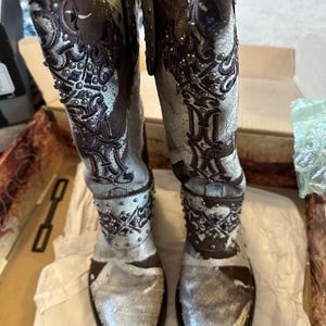 Può includere: Un paio di stivali alti in pelle invecchiata con punta. Gli stivali presentano ricami intricati, borchie argentate e un mix di colori marrone e bianco sporco. Gli stivali sono in una scatola, pronti per essere indossati.